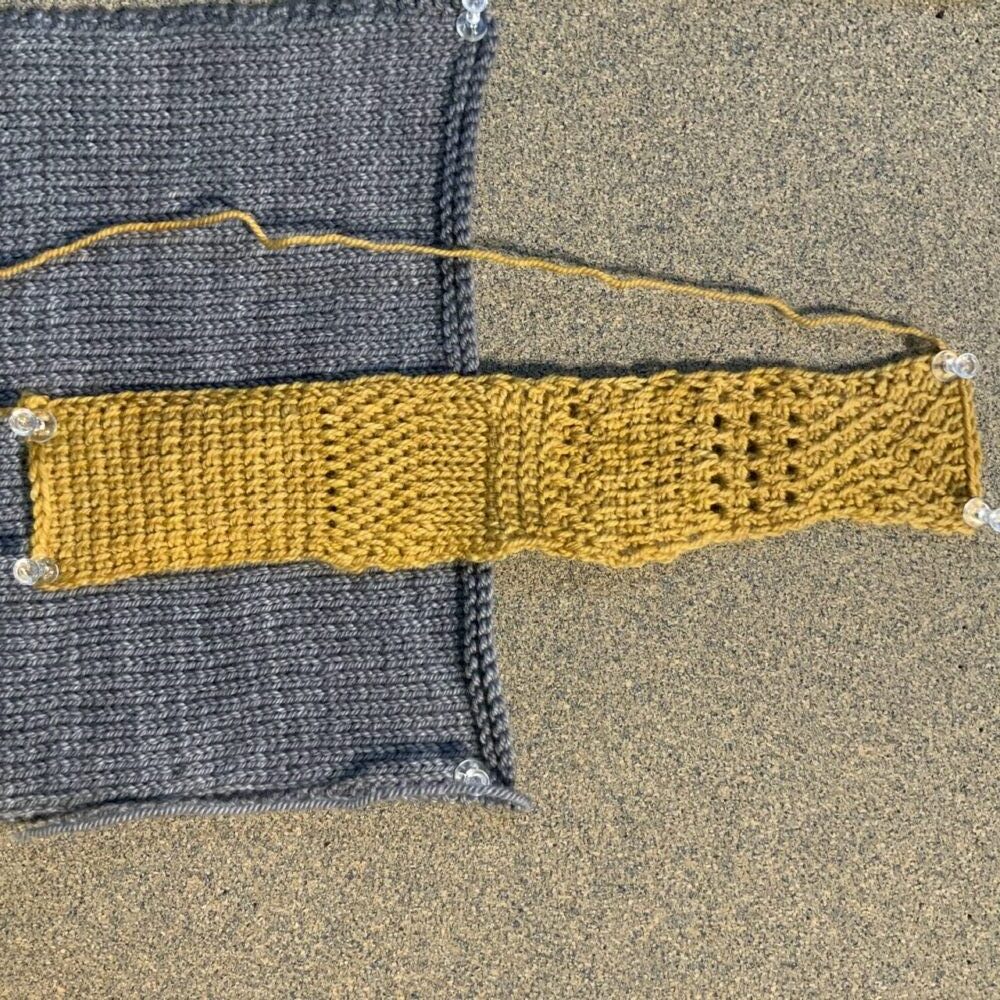 A gold strip of Tunisian crochet is pinned to a cork board. The strip displays several different Tunisian crochet stitches