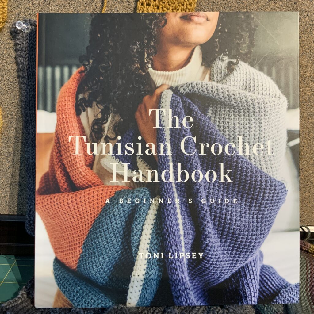 On the cover of the Tunisian Crochet Handbook by Toni Lipsey, a Black woman with long coiled hair wraps a colourful crocheted blanket around her.