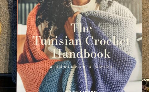 On the cover of the Tunisian Crochet Handbook by Toni Lipsey, a Black woman with long coiled hair wraps a colourful crocheted blanket around her.